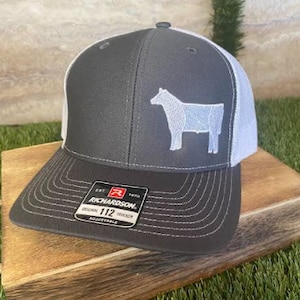 May include: A black and white trucker hat with a white silhouette of a cow on the front. The hat has a mesh back and a black adjustable strap with a red label that says "Richardson. Est. 1970. Original 112 Trucker. Adjustable."