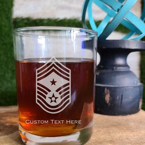 May include: A clear glass tumbler filled with a brown liquid, possibly whiskey or a similar beverage. The glass features a white military rank insignia and the text "CUSTOM TEXT HERE". The glass sits on a wooden surface.