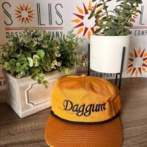 May include: A mustard yellow baseball cap with a structured crown and a flat brim. The cap features a black cord detail across the base of the crown and the word Daggum embroidered in black script lettering on the front center panel.