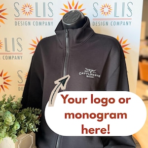 May include: A black full-zip jacket with a high collar and ribbed cuffs. The left chest features an embroidered white logo with a bull skull icon and the text "EST. 1910 CATTLEMENS BANK". A graphic overlay with an arrow points to the logo.