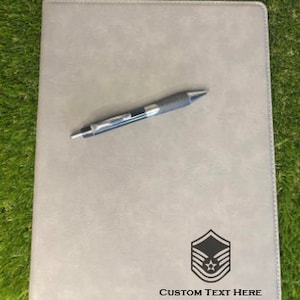 May include: A gray leatherette folder with a pen and a black and white Air Force insignia. The text "CUSTOM TEXT HERE" is printed below the insignia.
