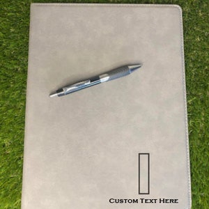 May include: A light gray rectangular portfolio folder with a smooth, matte finish and stitched edges. A retractable ballpoint pen with a dark gray grip and silver clip rests on the cover. The text "Custom Text Here" is printed in black.