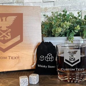 May include: A wooden box with a custom engraved design of an eagle, anchors, and chevrons. The box is accompanied by a glass with a similar design, a black bag with whiskey stones, and two slate coasters with the same design. The box and coasters have the text "CUSTOM TEXT" engraved on them.