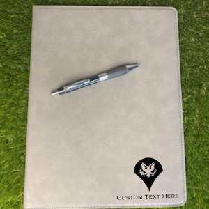 May include: A light gray notebook with a black pen resting on top. The notebook has a black emblem and the text "CUSTOM TEXT HERE" printed on the bottom right corner. The notebook is on a green surface.