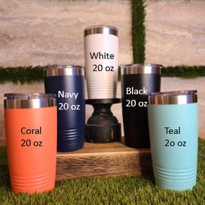 Op de afbeelding: Een verzameling van 20 oz ge&iuml;soleerde bekers in verschillende kleuren: koraal, marineblauw, wit, zwart en blauwgroen. Elke beker heeft een zilveren rand en een bijpassende deksel. De bekers worden op een houten oppervlak getoond.
