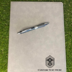 May include: A gray leatherette folder with a black pen and a black and white insignia with the text "CUSTOM TEXT HERE".