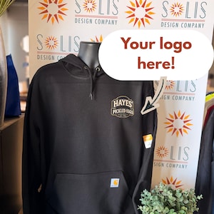 May include: A black hooded sweatshirt featuring a front kangaroo pocket and a small rectangular patch on the chest with the text Hayes Pickled Eggs Est. 2020. A small blue and yellow logo tag is visible on the lower pocket edge of the garment.