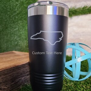 Op de afbeelding: Zwarte ge&iuml;soleerde beker met een zilveren rand en deksel. De beker heeft een witte omtrek van North Carolina en de tekst "Custom Text Here". De cilindrische beker is waarschijnlijk gemaakt van roestvrij staal.