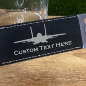 May include: A flat, stainless steel bottle opener featuring a dark blue, textured leather-like wrap secured with white stitching. The center displays a light gray silhouette of a fighter jet above the white text, CUSTOM TEXT HERE.