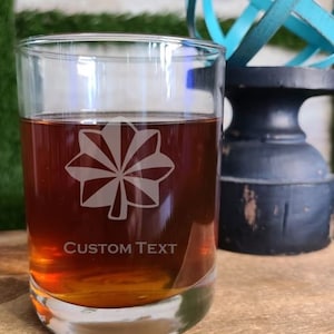 May include: A clear glass tumbler filled with a brown liquid, featuring a white etched floral design and the words "CUSTOM TEXT". The glass sits on a wooden surface, with a decorative blue metal sphere and black stand in the background.