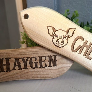 May include: Two wooden pig-themed brushes. One is light-colored with a pig face and the name "Chloe". The other is darker, with a pig silhouette in a barbed wire circle and the name "Haygen".