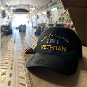 May include: A black baseball cap featuring yellow embroidered text that reads OPERATION EPIC FURY VETERAN. Above the word VETERAN is a small, rectangular, multi-colored ribbon bar graphic. The cap has a curved brim and a structured crown.