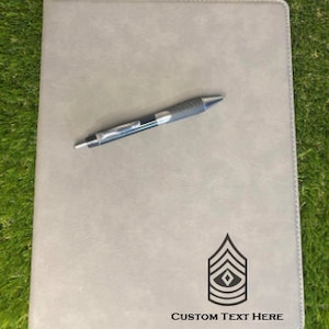 May include: A gray leatherette folder with a black pen and a black and white insignia. The text "CUSTOM TEXT HERE" is printed below the insignia.