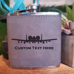 May include: A gray leather-wrapped flask with a silver top. The flask features a silhouette of an A-10 Warthog aircraft and the text "CUSTOM TEXT HERE".
