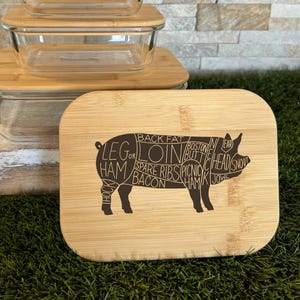 May include: A set of three rectangular glass food storage containers with light brown bamboo lids. The front-most lid features a dark brown laser-engraved diagram of a pig divided into various cuts of meat, including labels like ham, loin, and bacon.