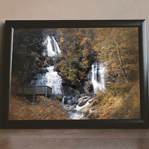 May include: A framed photograph of a waterfall cascading down a rocky cliffside, surrounded by lush green trees with vibrant autumn foliage. The water flows through a small wooden bridge in the foreground.