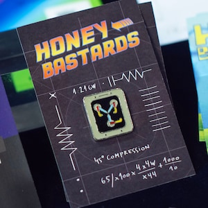 May include: A black pin featuring a silver frame with a black center and a stylized "Y" shape in red, yellow, and blue. The pin is on a black card with the text "HONEY BASTARDS" in orange and yellow, along with technical diagrams and equations.