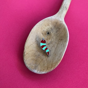 Peut inclure: Un petit charme émaillé rouge et vert en forme de piment, avec un contour argenté, est suspendu à un crochet argenté. Le charme repose sur une cuillère en bois sur un fond rose vif.