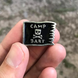 May include: A rectangular enamel pin featuring a black flag design with the words "CAMP BART" in white. The flag has a skull and crossbones emblem. The pin has a silver-colored border.