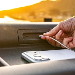 Peut inclure: Un smartphone blanc avec un câble de chargement connecté, posé sur une surface sombre. La scène est éclairée par la lumière chaude du soleil, suggérant un cadre extérieur. Le téléphone est à côté d'un port de chargement.