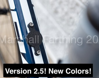 May include: A black bicycle accessory is mounted on a blue and white bicycle frame. The accessory is round with two mounting points. Text at the bottom reads "Version 2.5! New Colors!" The image also includes the text "© Marshall Farthing 2021".