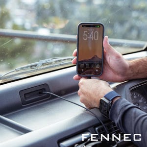May include: A smartphone in a blue case is connected to a charging cable inside a vehicle. The phone screen shows a mountain scene and the time 5:40. A smartwatch with a blue strap is on the wrist. The word "FENNEC" is at the bottom.