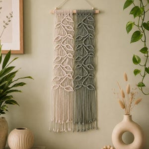 May include: A macrame wall hanging featuring two vertical sections of knotted cotton cord in cream and sage green. The design is suspended from a horizontal wooden dowel with a thin cord loop for hanging, ending in loose, knotted fringe at the base.