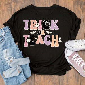 May include: Black t-shirt with the text "Trick or Teach" in pink, orange, and purple with a ghost, bat, and pumpkin graphic. The shirt is laying on a wooden surface with blue jeans and a pair of white and pink sneakers.