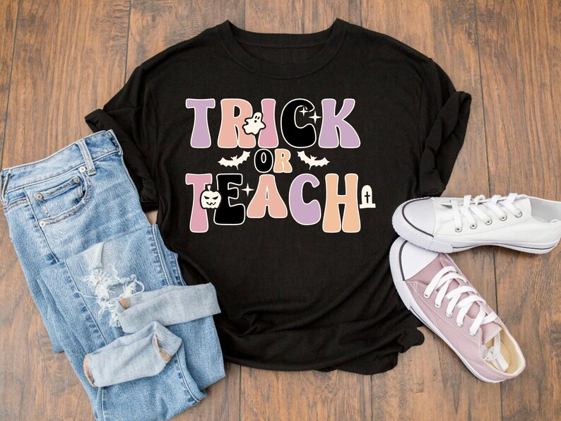 May include: Black t-shirt with the text "Trick or Teach" in pink, orange, and purple with a ghost, bat, and pumpkin graphic. The shirt is laying on a wooden surface with blue jeans and a pair of white and pink sneakers.
