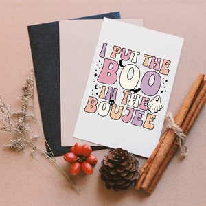 Puede incluir: Una tarjeta blanca con el texto "I put the BOO in the BOUJEE" en letras de colores. La tarjeta est&aacute; rodeada de un fondo marr&oacute;n con palitos de canela y una pi&ntilde;a.