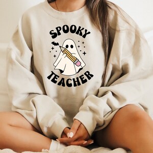 May include: A beige sweatshirt with the text "Spooky Teacher" in black lettering. A cartoon ghost is holding a pencil in front of the text.