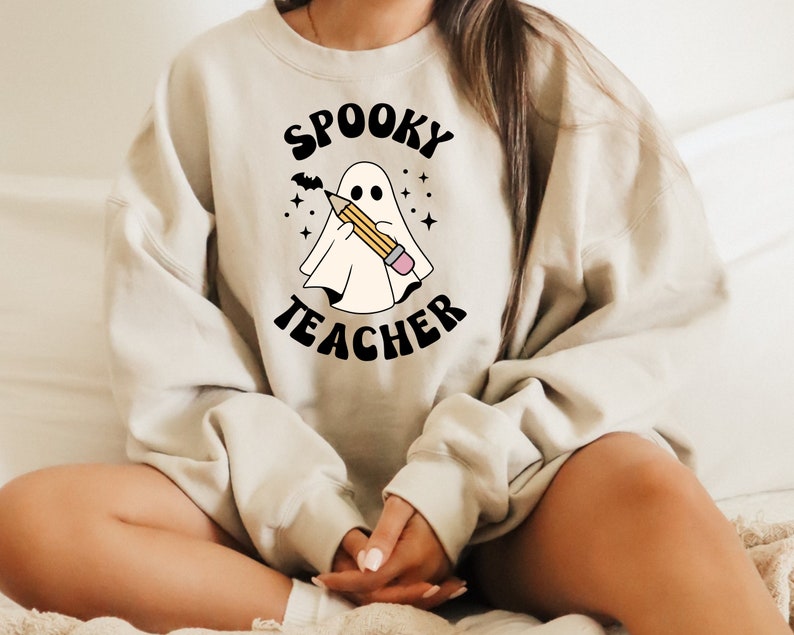 May include: A beige sweatshirt with the text "Spooky Teacher" in black lettering. A cartoon ghost is holding a pencil in front of the text.