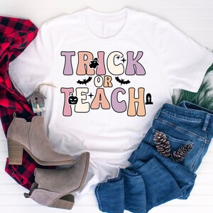 May include: White t-shirt with the text "Trick or Teach" in colorful retro letters. The letters are decorated with Halloween icons like ghosts, bats, and pumpkins.