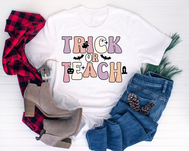 May include: White t-shirt with the text "Trick or Teach" in colorful retro letters. The letters are decorated with Halloween icons like ghosts, bats, and pumpkins.