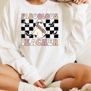 May include: White sweatshirt with a black and white checkered background, a ghost holding a pencil, and the text "FABOOLOUS TEACHER" in pink and orange letters.