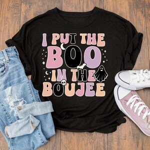 Puede incluir: Camiseta negra con un estampado gr&aacute;fico que dice "I put the boo in the boujee" en letras rosa, morado y naranja. El gr&aacute;fico tambi&eacute;n incluye un fantasma, un murci&eacute;lago y una calavera.