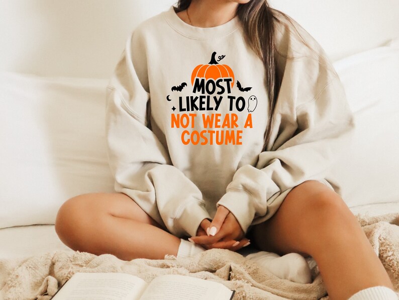 May include: A light beige sweatshirt with a pumpkin graphic and the text "Most Likely To Not Wear A Costume".