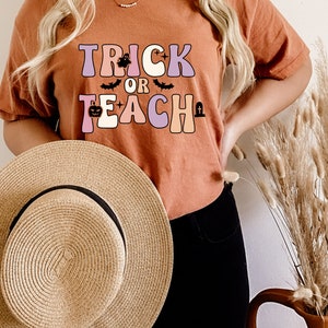May include: A woman wearing a rust-colored t-shirt with the text "Trick or Teach" in pastel colors. The text is decorated with a ghost, bats, and a pumpkin. She is holding a straw hat in front of her.
