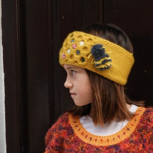 May include: Mustard yellow knitted headband with a bee embellishment and floral accents. The headband is worn by a person wearing a colourful sweater with orange and red tones. The headband has a textured design.