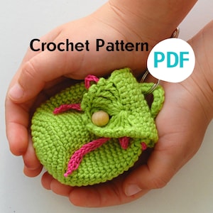 Può includere: Una pochette verde all'uncinetto con rifiniture rosa e un bottone di legno. La pochette è tenuta nelle mani di una persona. Il testo "Crochet Pattern PDF" è visibile nell'immagine.