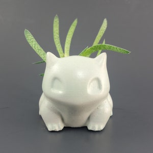 May include: A light grey concrete planter in the shape of a cartoon character, with a small green plant growing from the top. The planter has a smooth, matte finish and is on a dark grey surface.