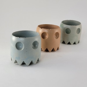 Puede incluir: Tres pequeñas macetas de hormigón con forma de fantasma, dispuestas en fila. Las macetas son de color azul, beige y verde. Cada una tiene dos agujeros redondos para los ojos y un borde inferior dentado. Ideales para plantas pequeñas.