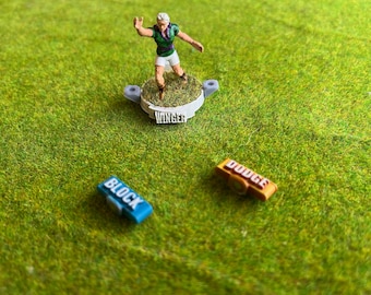 May include: A miniature figurine of a player in green and purple, standing on a base marked "WINGER." Two game markers, labelled "BLOCK" and "DODGE", are also present. The scene is set on a green, grassy surface.