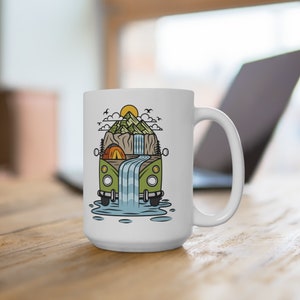 May include: White ceramic mug with a green and brown illustration of a vintage camper van driving through a waterfall in a mountain landscape. The camper van has a yellow tent inside.