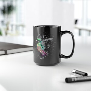 May include: Black ceramic mug with a white astronaut riding a colorful jellyfish in space. The mug has a black handle and a white rim.