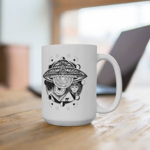 May include: White ceramic mug with a black and white illustration of a UFO, an eye, pyramids, and a woman's face. The design is surrounded by a circle of moons and stars.