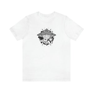 May include: White t-shirt with a black and white graphic print of a UFO with a face inside. The UFO is surrounded by a circle of stars and moons.