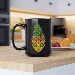 May include: Black ceramic mug featuring a whimsical design of a pineapple with an alien face. The pineapple has a yellow and orange exterior with green leaves. The alien has green eyes and a smiling expression. The mug has a large handle.