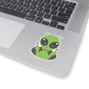 May include: A green alien sticker with large black eyes and small red hearts. The alien is making a heart shape with its fingers. The sticker is on a laptop.