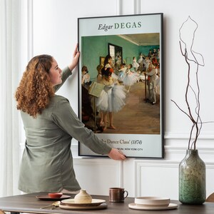Pu&ograve; includere: Una donna appende una stampa d'arte incorniciata di "La Classe di Danza" (1874) di Edgar Degas su una parete bianca. La stampa presenta un gruppo di ballerine in uno studio di danza, con una parete verde chiaro e un pavimento in legno.